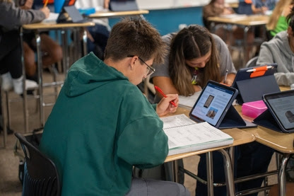Un estudiante con una sudadera con capucha verde toma apuntes mientras estudia en un pupitre; al fondo se ven sus compañeros de clase y sus tabletas.