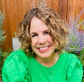A person with curly hair smiles, wearing a bright green top, in a garden with a wooden fence and purple flowers.